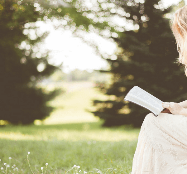 Frau sitzt auf einem Stein mittten in einer grünen Wiese mit Bäumen im Hintergrund und liest ein Buch
