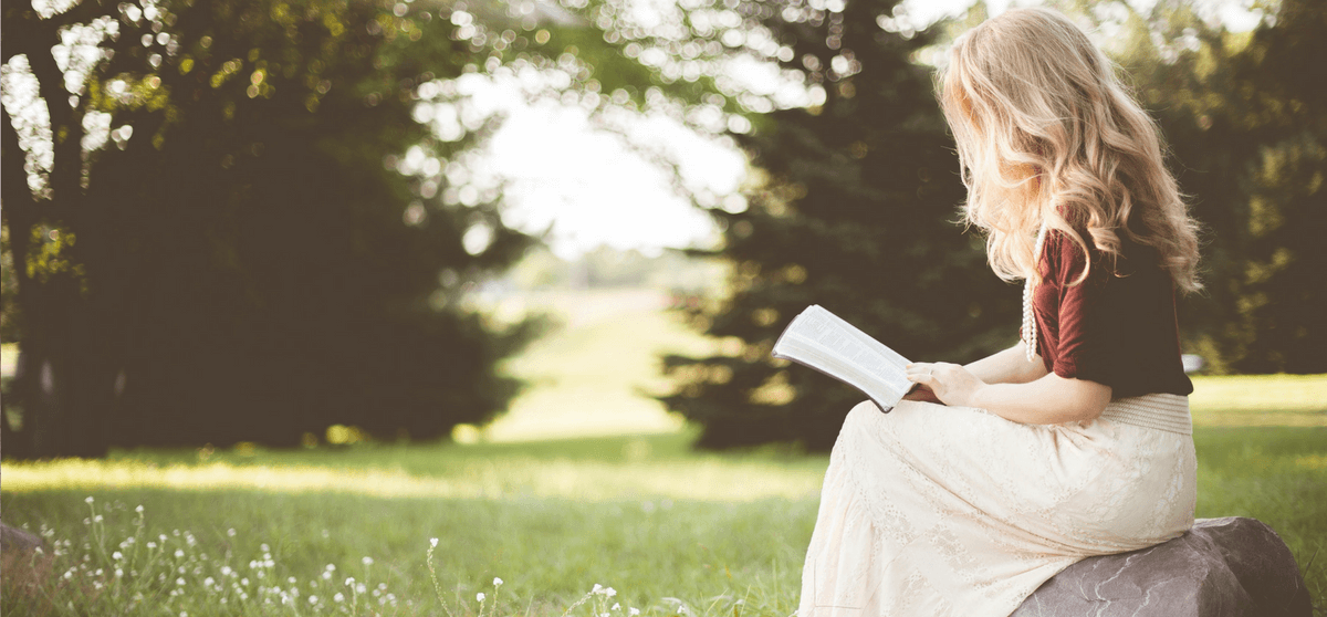 Frau sitzt auf einem Stein mittten in einer grünen Wiese mit Bäumen im Hintergrund und liest ein Buch