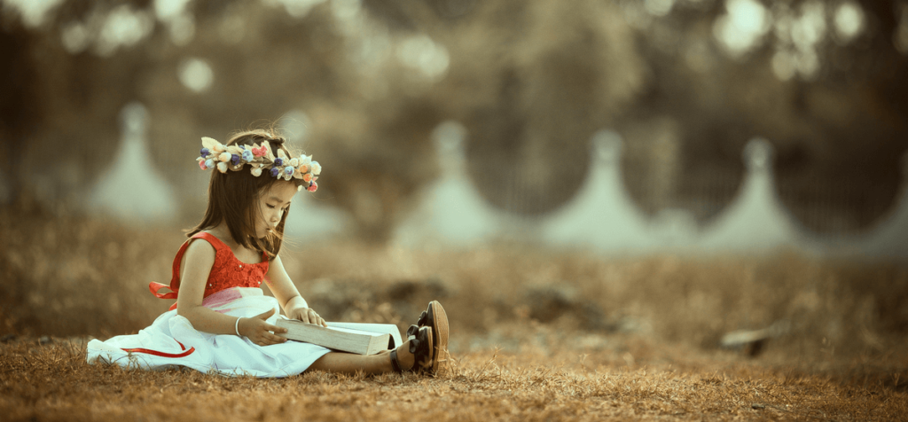 Mädchen mit Blumenkranz im Haar liest ein Buch und sitzt auf einer Wiese